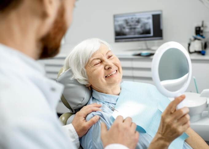 Senior dental patient admiring her smile in mirror after replacing missing teeth in Orem