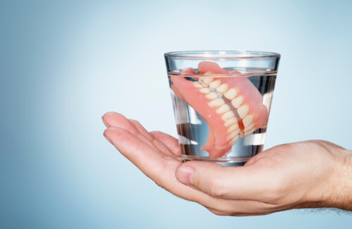 Hand holding a glass of water with a set of full dentures soaking inside of it