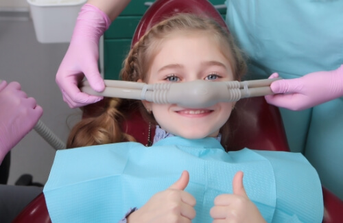 Young girl in dental chair smiling and giving thumbs up with nitrous oxide mask on her nose